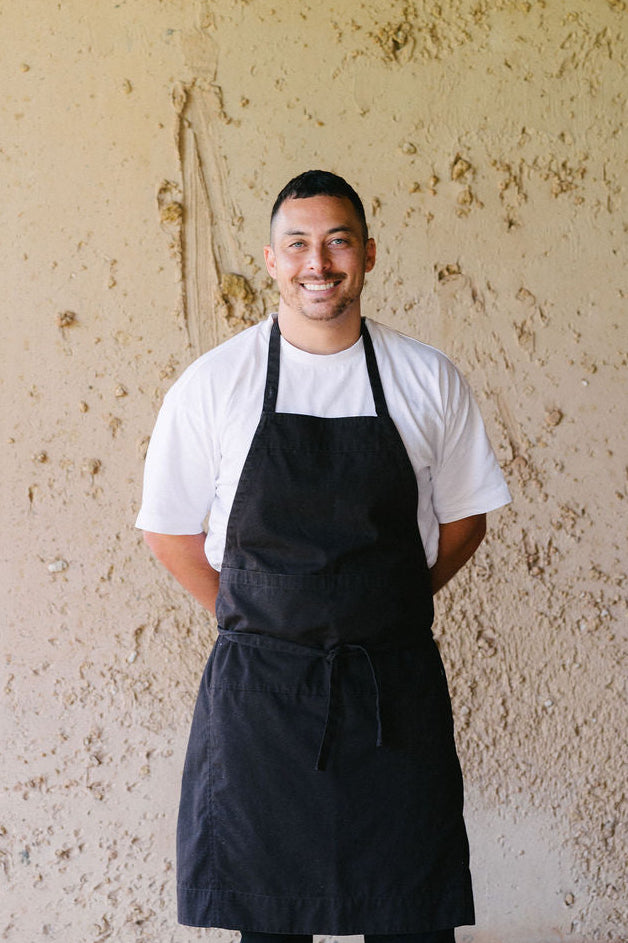 Head Chef wearing a black apron over a white shirt against a textured beige wall.