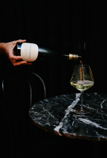 Person pouring white wine from a bottle into a glass on a dark marble table.