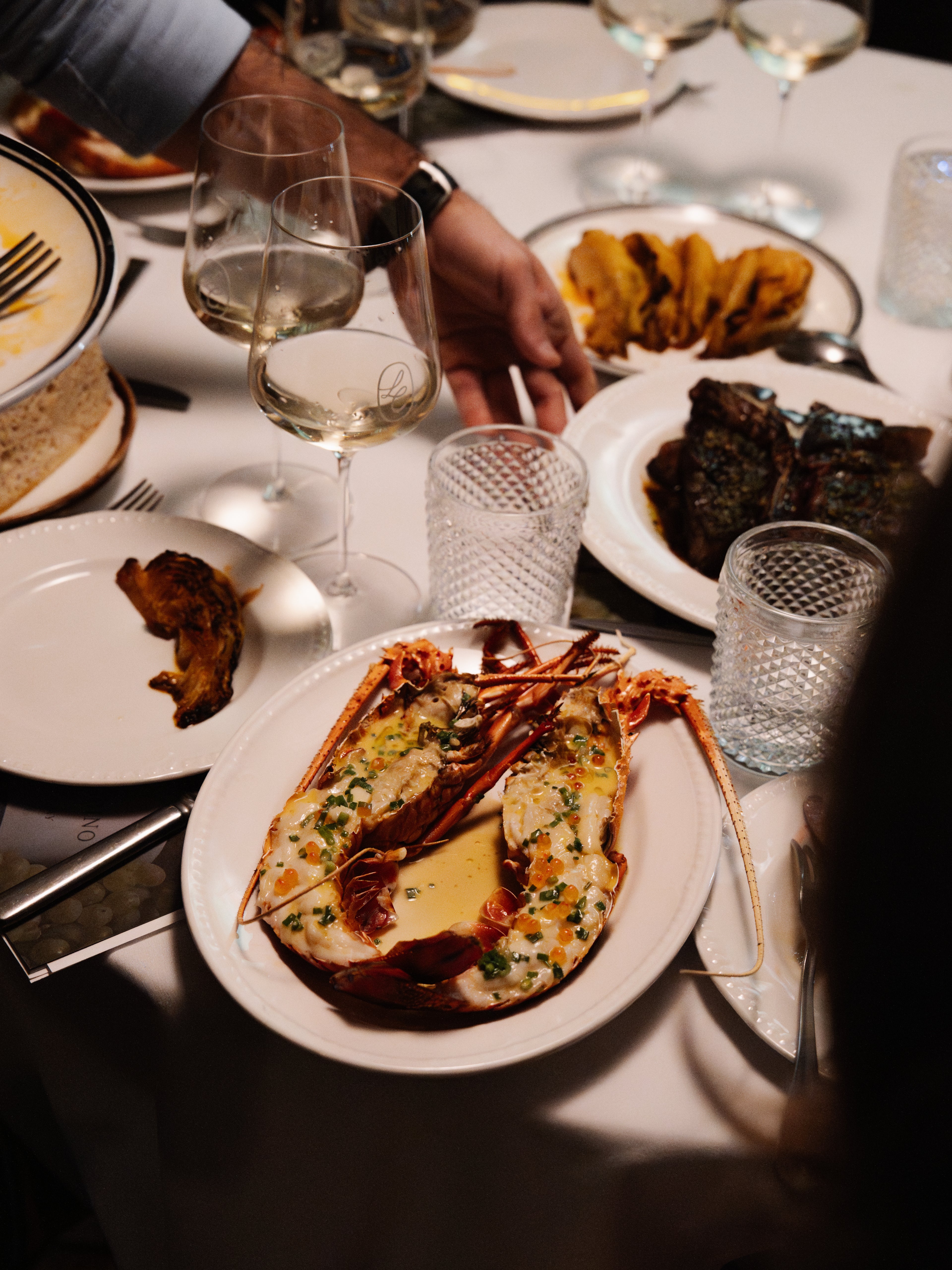 Dinner table with plates of food, glasses, and a person's hand holding a glass.
