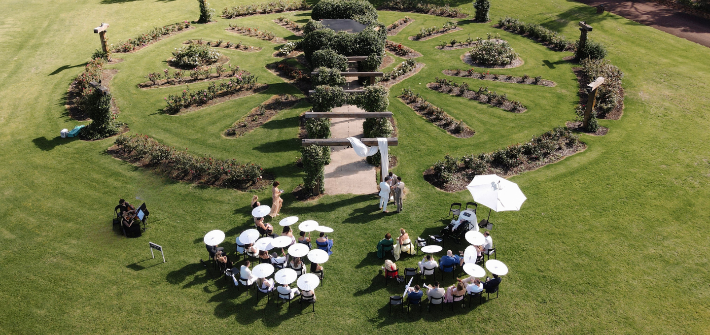 A wedding in a formal garden with people under white umbrellas, surrounded by well-manicured lawns, rose gardens and pathways.