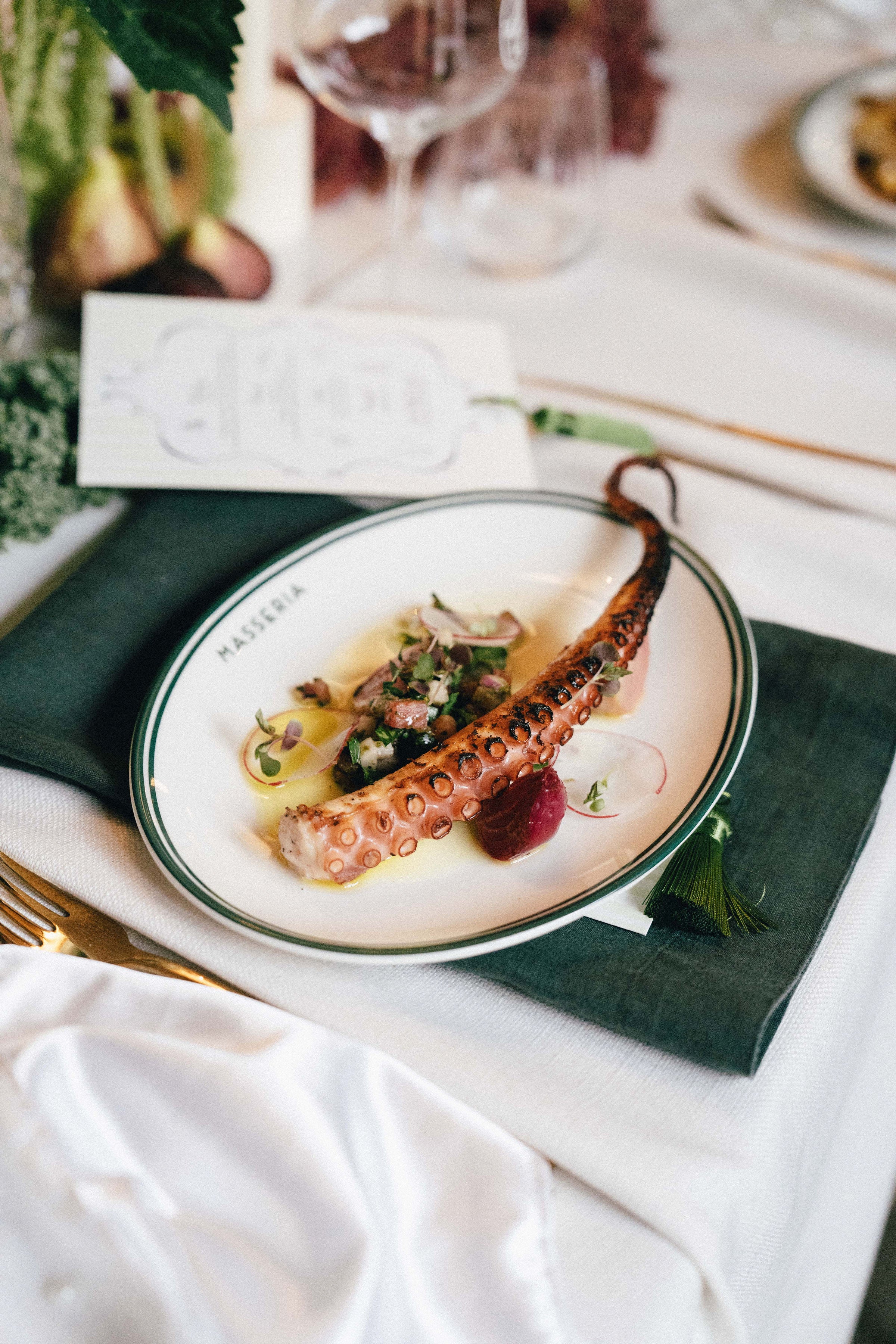 Gourmet dish with octopus on a plate, served on a white tablecloth with green napkins.