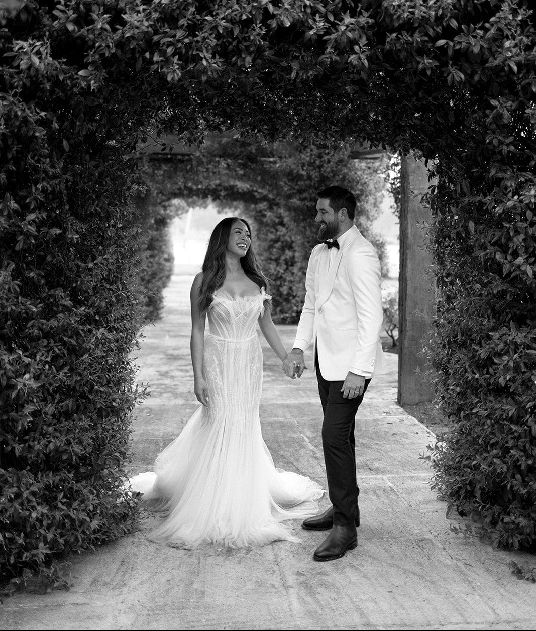 Black and white photo of a bride and groom standing under an archway in a garden.