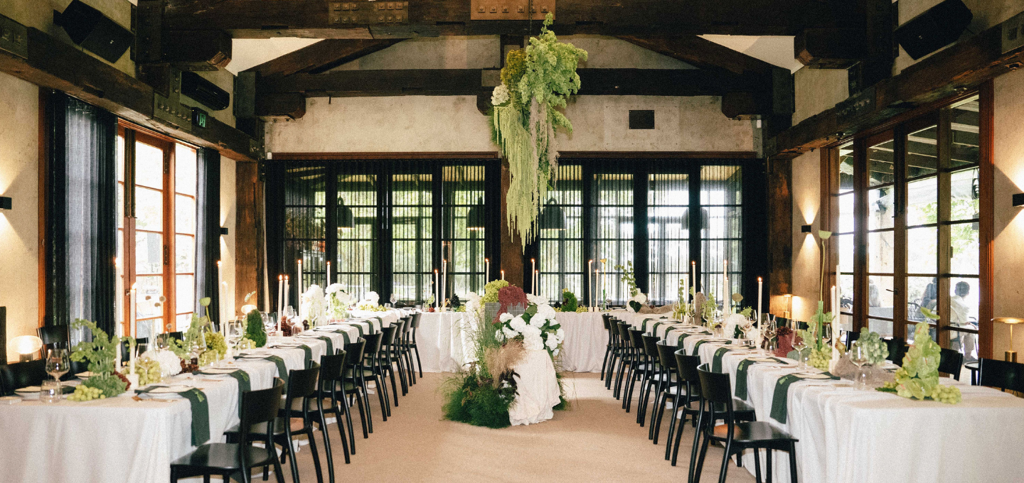 Decorated dining room with tables set for a wedding, large windows, and wooden beams.