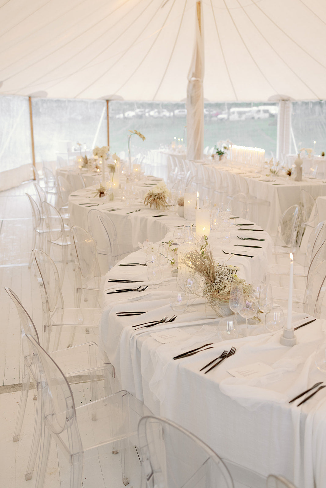 Elegant white table setting with clear chairs and floral centerpieces in a tented outdoor area.