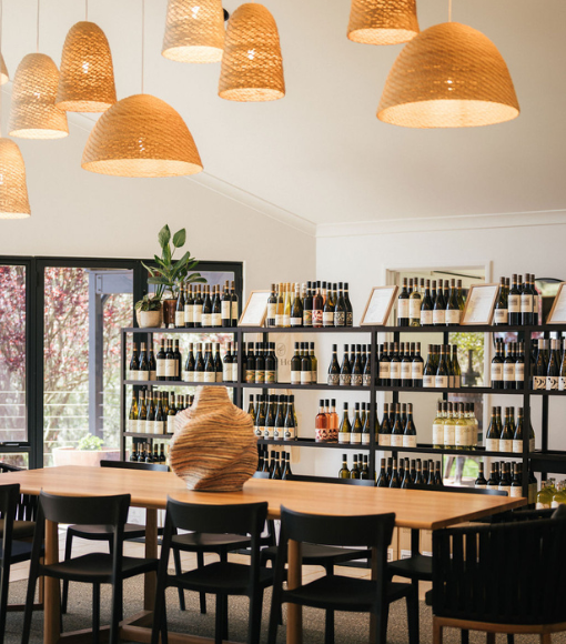 Ad Hoc Cellar Door. Wine store interior with wooden table, chairs, and shelves stocked with wine bottles.