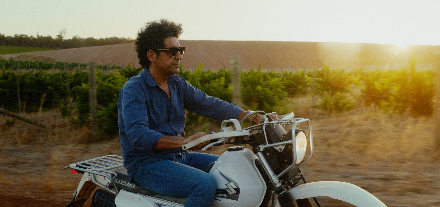 Larry Cherubino riding a motorcycle in a vineyard and sunset in the background